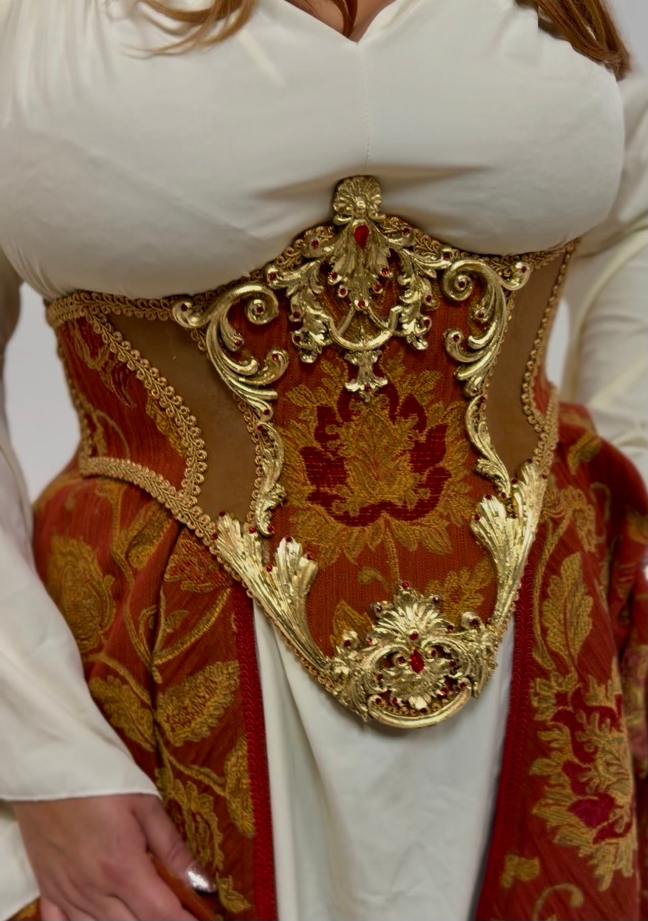 A close-up of a person wearing an ornate medieval-style corset with gold and red brocade detailing over a white dress.